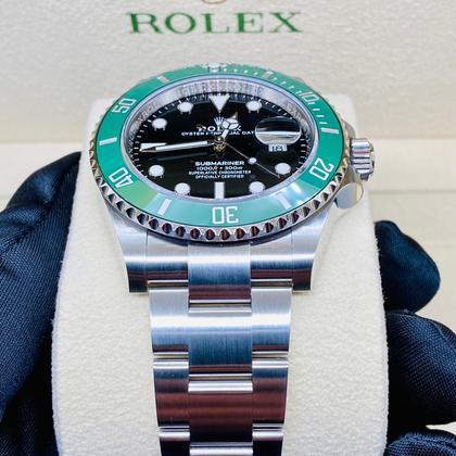 Rolex watch with green bezel and black face on a white cushion, with 'Rolex' branding in the background.
